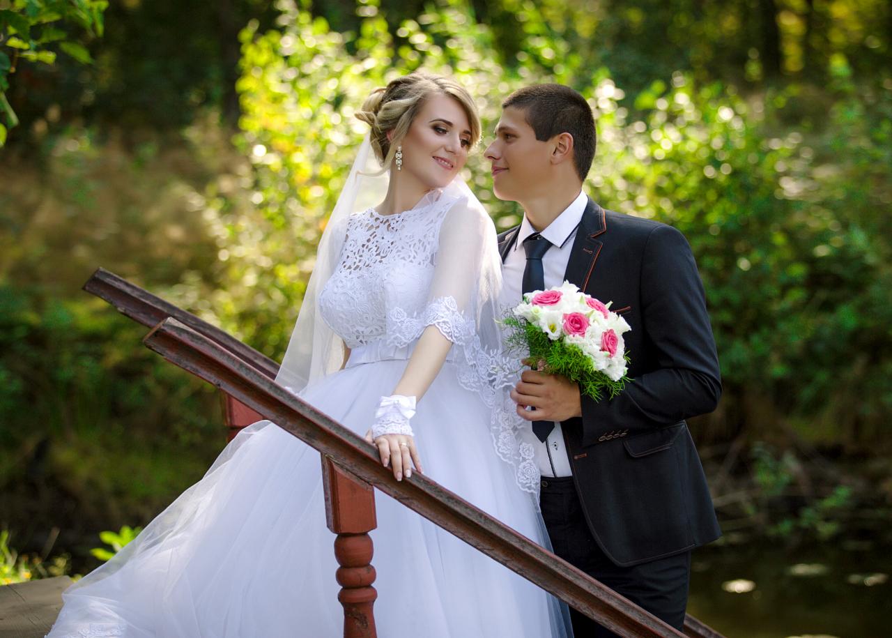 Para młoda na drewnianym mostku w lesie, panna młoda w koronkowej sukni i z welonem, pan młody w czarnym garniturze z bukietem białych i różowych róż.