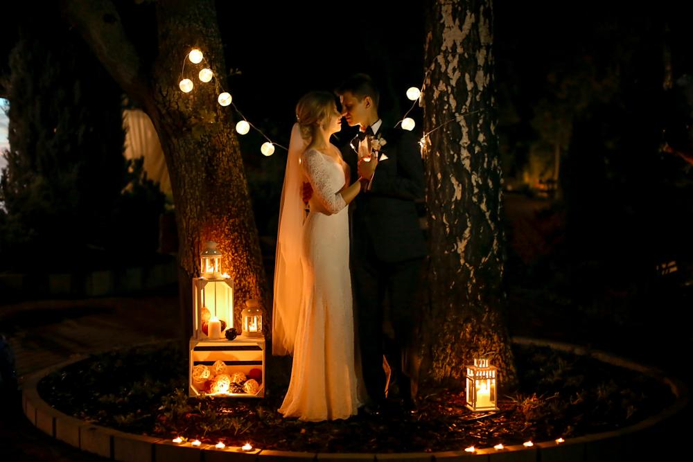 Para młoda w nocnej scenerii, otoczona drzewami z girlandami świetlnymi, lampionami i świecami, tworząc romantyczny nastrój.