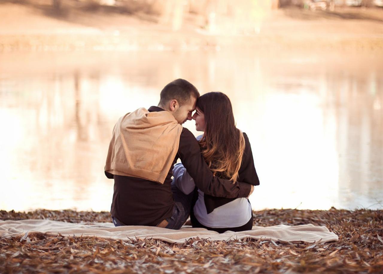 Zakochana para siedzi tyłem na kocu nad brzegiem jeziora, dotykając się czołami w intymnej chwili.