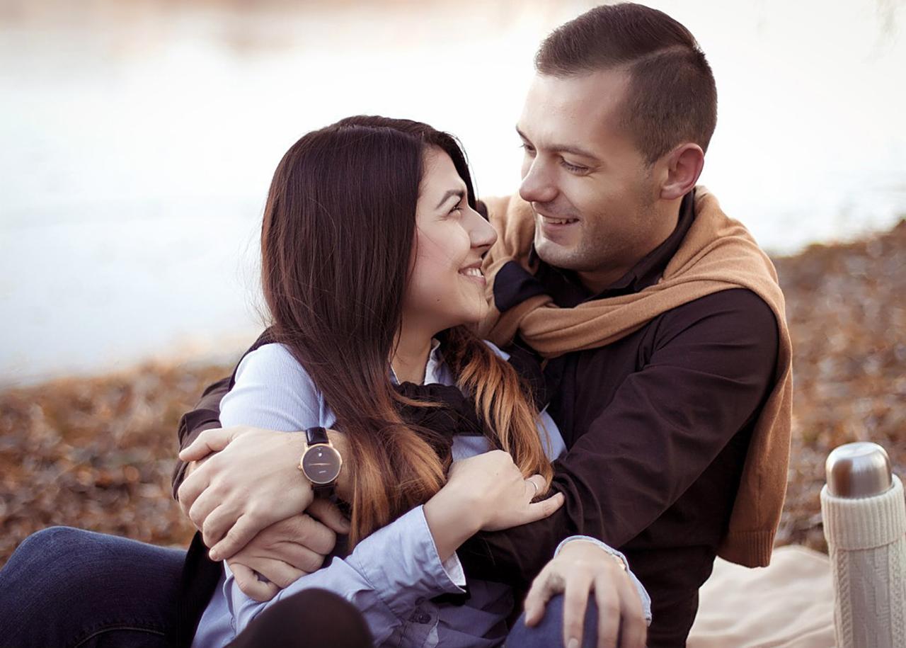 Uśmiechnięta para zakochanych siedzi blisko siebie na tle jesiennego krajobrazu; mężczyzna obejmuje kobietę, oboje patrzą na siebie z czułością. Na pierwszym planie widoczny damski zegarek na ręce.