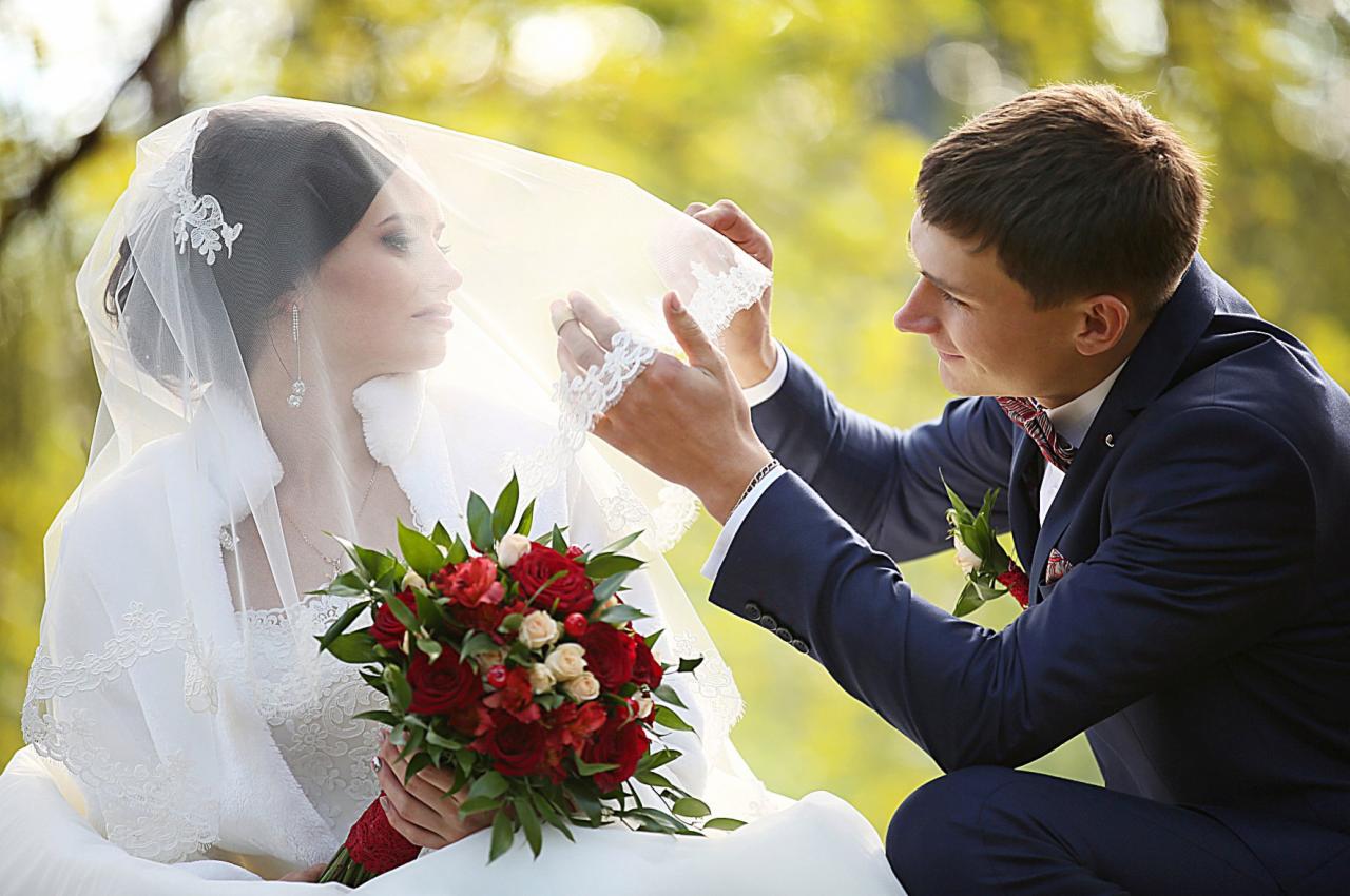 Panna młoda z bukietem czerwonych róż, spogląda spod welonu, który poprawia pan młody w granatowym garniturze z bordową muszką.