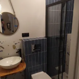 Nowoczesna łazienka z umywalką nablatową na drewnianym blacie, wiszącą miską WC obudowaną niebieskimi płytkami oraz kabiną prysznicową z czarnymi profilami i niebieskimi płytkami, uzupełniona...