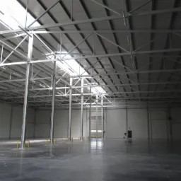 Wnętrze hali stalowej z widoczną konstrukcją nośną dachu, szara betonowa posadzka z żółtymi zabezpieczeniami słupów, światło dzienne wpadające przez świetliki dachowe.