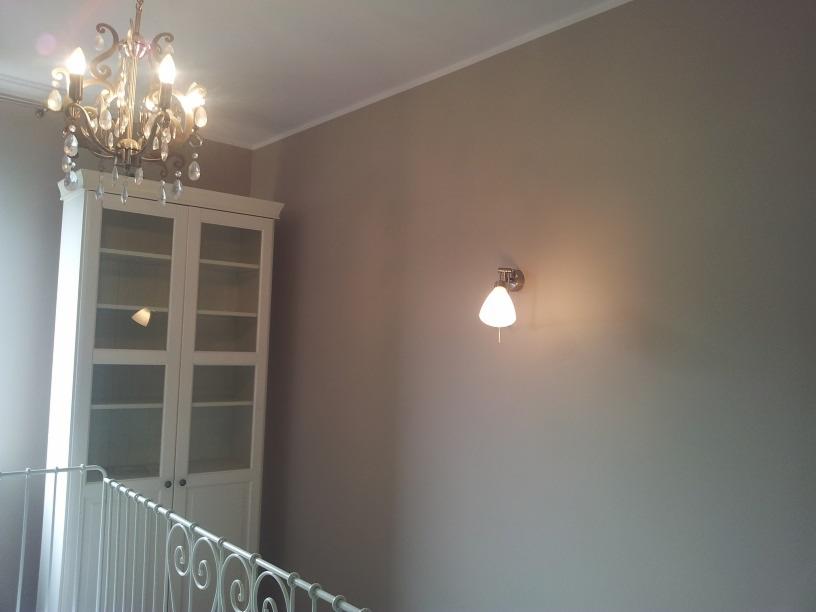 Biała, przeszklona witryna z oświetleniem wewnętrznym, widoczna spod balustrady schodów, z ozdobnym żyrandolem i kinkietem na szarej ścianie.