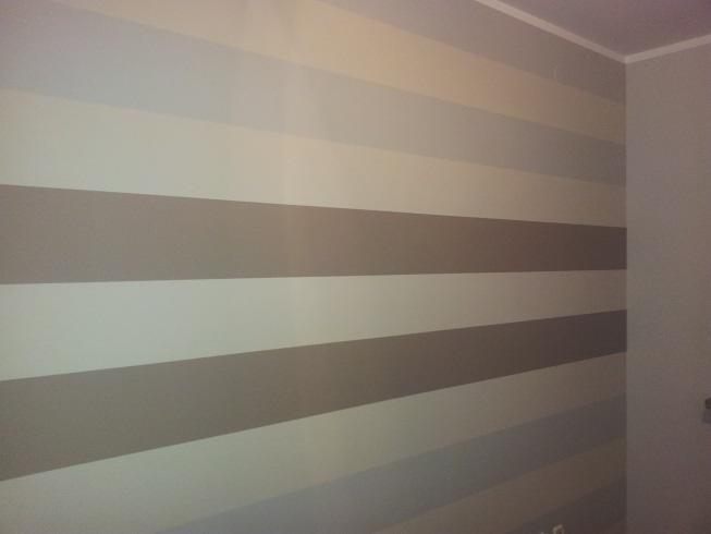 Ściana pomalowana w poziome pasy w odcieniach szarości i beżu, widoczny fragment sufitu i narożnik pokoju.