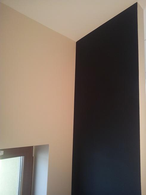 Fragment pokoju z kontrastującymi ścianami: beżowa i czarna, z widocznym fragmentem okna z brązową ramą w dolnym rogu.