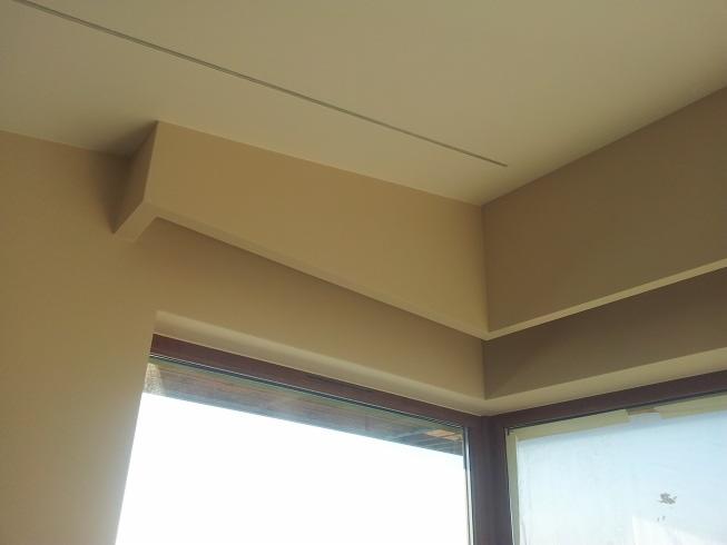 Beżowe ściany i sufit połączone gładzią, widoczne okno z brązową ramą i fragment szyny oświetleniowej na suficie.