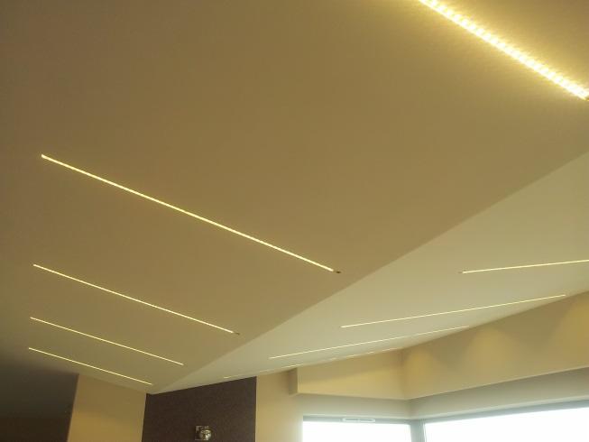 Nowoczesny sufit z wbudowanymi liniowymi oprawami LED, tworzącymi geometryczny wzór, oraz dodatkowe oświetlenie LED wzdłuż krawędzi sufitu podwieszanego.