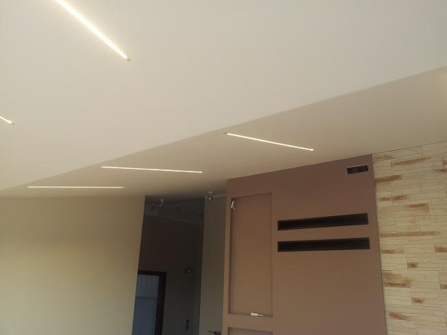 Nowoczesne oświetlenie sufitowe z liniowymi lampami LED w minimalistycznym wnętrzu, z akcentem na ścianę z dekoracyjną okładziną przypominającą piaskowiec.