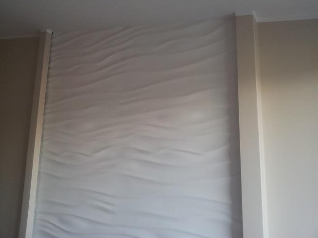 Ściana z dekoracyjną, trójwymiarową strukturą fal, obramowana pionowymi listwami w odcieniu złamanej bieli, przylegająca do gładkiej, beżowej ściany.