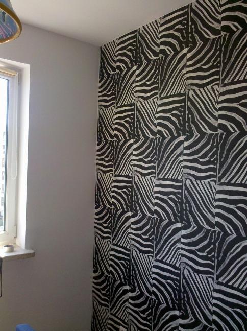 Ściana w pokoju wykończona tapetą w motyw zebry, sąsiadująca z szarą ścianą i oknem z białymi ramami.