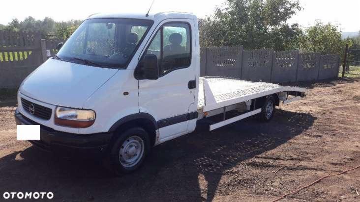 Biała laweta Opel Movano z płaską platformą, zaparkowana na ziemnym placu w słoneczny dzień, widoczny fragment ogrodzenia z betonowych płyt w tle.