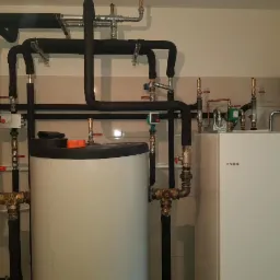 Kotłownia z zasobnikiem ciepłej wody i piecem NIBE, liczne rury, zawory i osprzęt w schludnym pomieszczeniu.