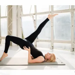 Kobieta w czarnym stroju wykonuje ćwiczenie na macie, unosząc jedną nogę w górę, w tle okna z kratami.