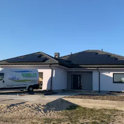 Dom jednorodzinny z panelami słonecznymi na dachu, na podjeździe zaparkowany biały bus z logo firmy instalującej panele, teren wok&oacute;ł domu w trakcie prac wykończeniowych.