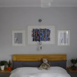 Jasnoszara sypialnia z drewnianym łóżkiem, trzema obrazami w białych ramach nad wezgłowiem, lampą wiszącą i pluszowym misiem na pościeli.