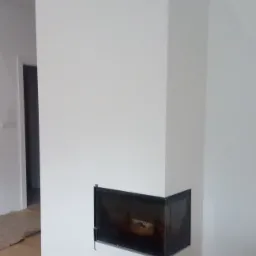 Biała obudowa kominka z czarnymi akcentami w nowo wykończonym wnętrzu, widoczna podłoga z paneli i fragment ściany po lewej stronie.