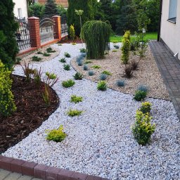 Ogrody Gniezno Łukasz Kaczała - Ogród z białym żwirem, ozdobnymi krzewami i korą drzewną, obrzeża z kostki brukowej. Widok na ogrodzenie z cegły i metalu oraz fragment elewacji budynku.