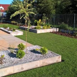 Ogrody Gniezno Łukasz Kaczała - Nowoczesny ogród z równo przystrzyżonym trawnikiem, betonowymi rabatami wypełnionymi kamieniami i roślinami, oraz ścieżką z płyt. W tle dom z czerwoną dachówką.