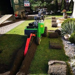 Ogrody Gniezno Łukasz Kaczała - Zielony traktor wykopuje rów pod trawnik z rolki. Widoczne fragmenty trawy z rolki, przyczepa i drzewa w tle. Praca w toku, świeżo wykopany rów.