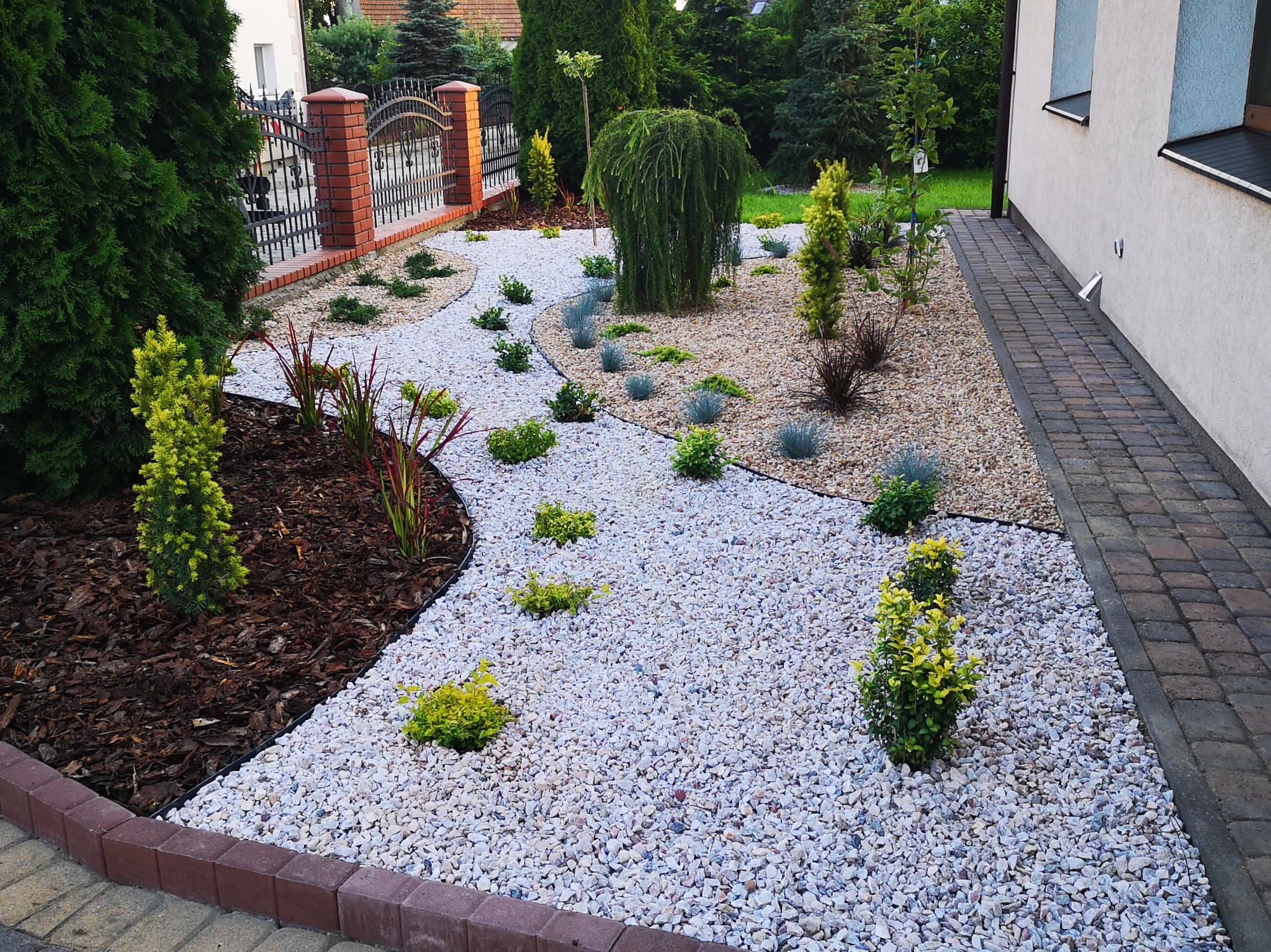 Ogród z białym żwirem, ozdobnymi krzewami i korą drzewną, obrzeża z kostki brukowej. Widok na ogrodzenie z cegły i metalu oraz fragment elewacji budynku.