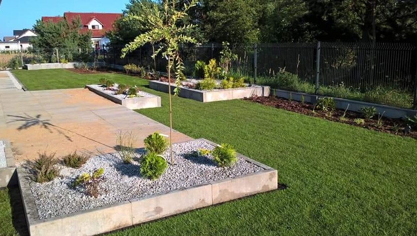 Nowoczesny ogród z równo przystrzyżonym trawnikiem, betonowymi rabatami wypełnionymi kamieniami i roślinami, oraz ścieżką z płyt. W tle dom z czerwoną dachówką.