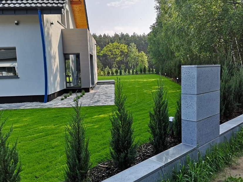 Nowoczesny ogród z równo przystrzyżonym trawnikiem, ścieżką z kostki i niskim murem z betonowych elementów. W tle widoczny dom i drzewa.