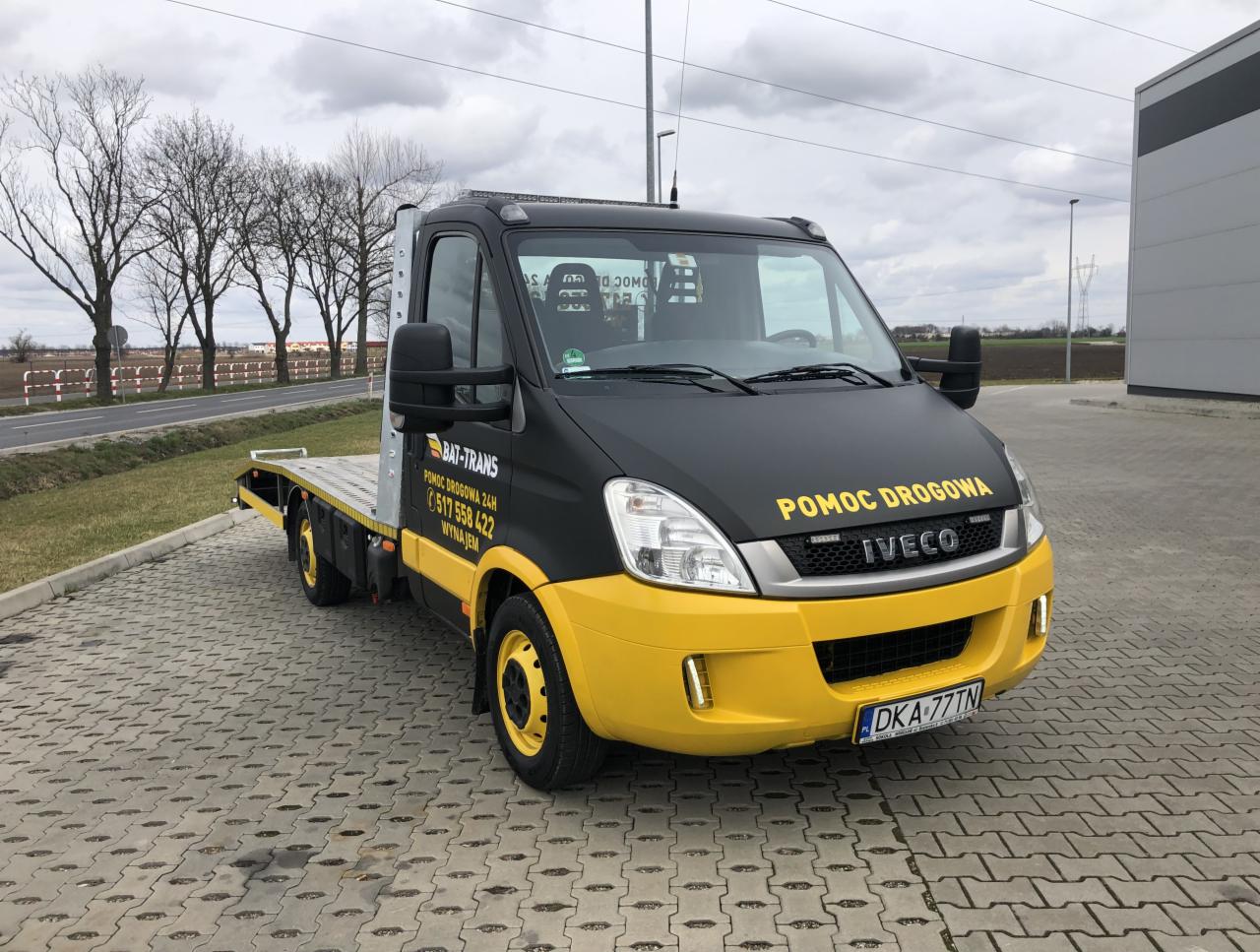 Czarno-żółta laweta marki Iveco z napisem 'Pomoc Drogowa' na parkingu z kostki brukowej, widok z przodu na tle drzew i budynku.