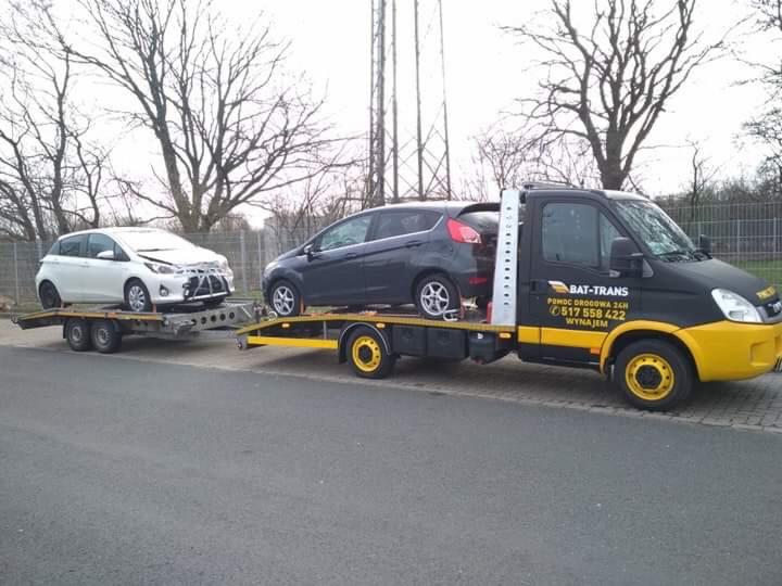 Czarno-żółta laweta z logiem BAT-TRANS przewozi dwa samochody osobowe: biały uszkodzony i grafitowy, na tle drzew bez liści.
