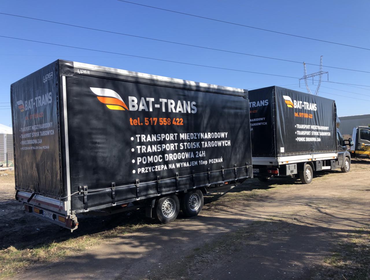 Dwa busy z przyczepami firmy Bat-Trans z czarnymi plandekami, na których widnieje logo i numer telefonu, zaparkowane na placu z widoczną linią wysokiego napięcia w tle.