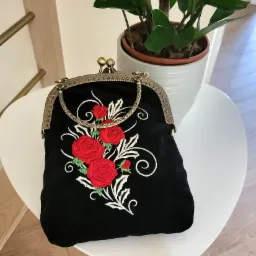 Czarna, aksamitna torebka z metalowym zapięciem i haftem przedstawiającym czerwone róże z białymi ornamentami, ustawiona na białym stoliku obok doniczki z zieloną rośliną.