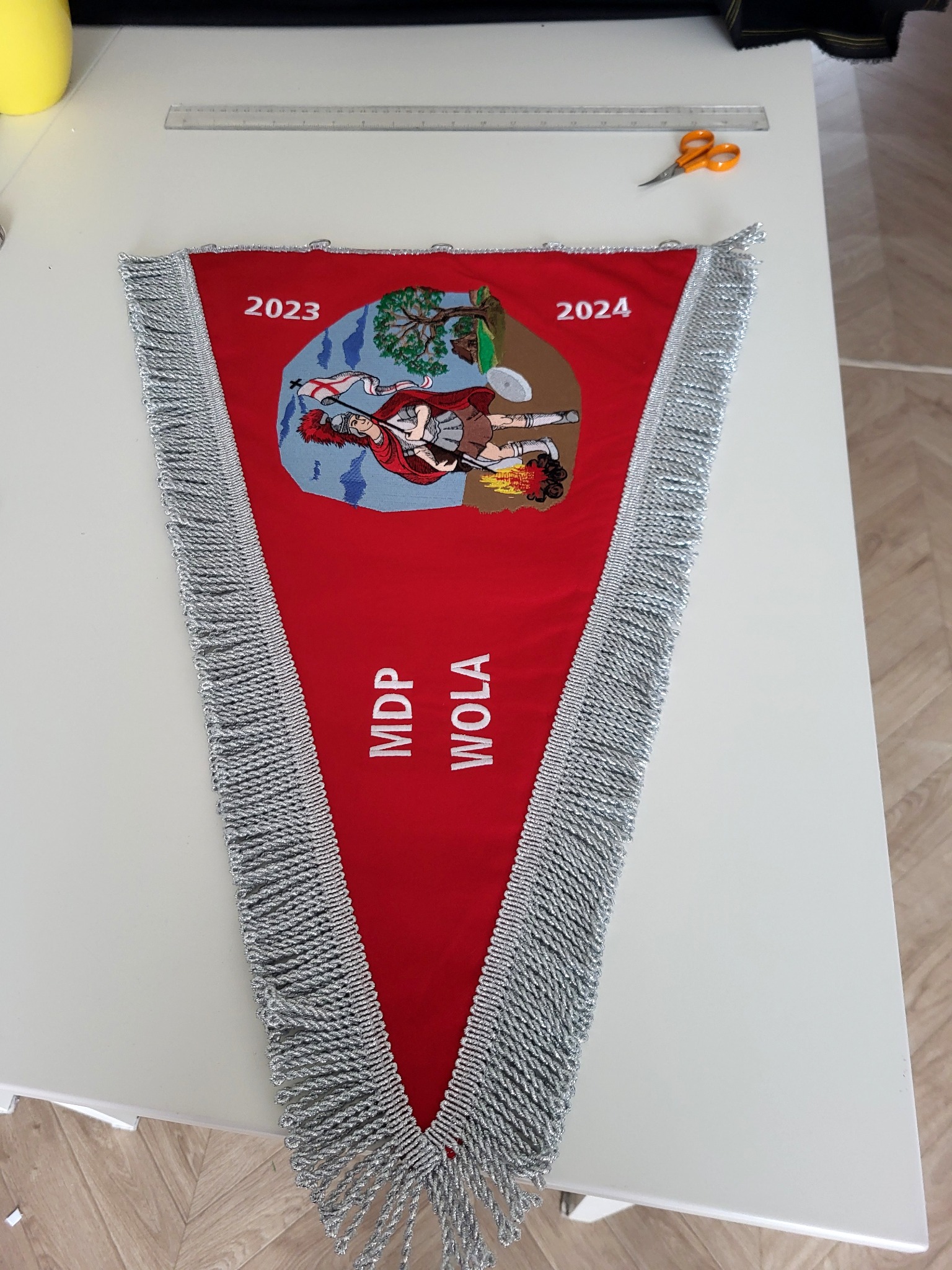 Czerwony trójkątny proporzec z srebrnymi frędzlami, haftowanym herbem, datami 2023 i 2024 oraz napisem 'MDP WOLA', leżący na białym stole obok linijki i nożyczek.