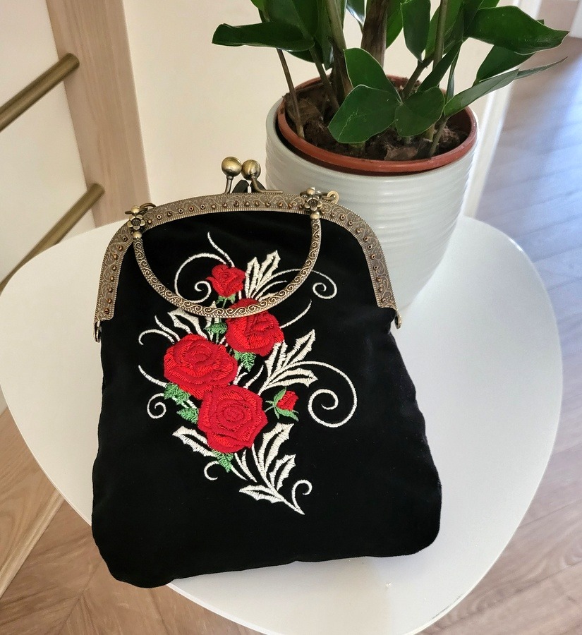 Czarna, aksamitna torebka z metalowym zapięciem i haftem przedstawiającym czerwone róże z białymi ornamentami, ustawiona na białym stoliku obok doniczki z zieloną rośliną.