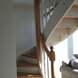 Drewniane schody z białymi balustradami i drewnianą poręczą, oświetlone naturalnym światłem, widoczne z dołu, z fragmentem szarej sofy w tle.