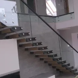 Nowoczesne, samonośne schody z drewnianymi stopniami i szklaną balustradą mocowaną na metalowych elementach, w jasnym wnętrzu domu.