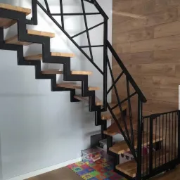 Nowoczesne schody z drewnianymi stopniami i czarną, geometryczną konstrukcją metalową, zabezpieczone barierką ochronną na dole, na tle drewnianej ściany i szarej ściany, z widoczną kolorową matą...