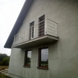 Szary dom jednorodzinny z balkonem zabezpieczonym prostą, metalową balustradą z poziomych prętów, widok z dołu na tle pochmurnego nieba.