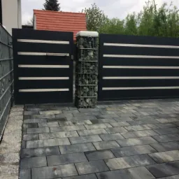 Nowoczesna, przesuwna brama wjazdowa w kolorze antracytowym z poziomymi, srebrnymi panelami, wsparta na słupku z gabionów wypełnionych kamieniami, kostka brukowa na podjeździe.