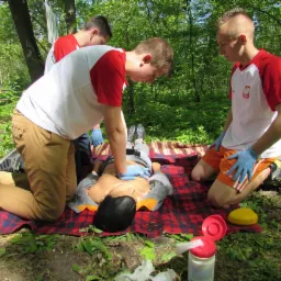 Instruktorzy demonstrujący resuscytację krążeniowo-oddechową na fantomie podczas szkolenia na świeżym powietrzu, widoczne rękawice ochronne i akcesoria do ćwiczeń na tle leśnym.