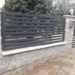 Nowoczesne, poziome przęsła ogrodzeniowe w kolorze grafitowym, zamontowane na słupkach i podmurówce z szarej kostki łupanej. Widoczna furtka z tym samym wypełnieniem. W tle zieleń drzew i krzewów.