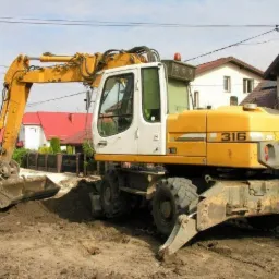 Żółto-biała koparka kołowa Liebherr 316 w trakcie prac ziemnych na tle domów jednorodzinnych, widoczne ślady świeżo wykopanej ziemi.