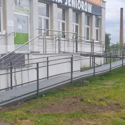 IMAGINATIVE Robert Sendrowicz - Zewnętrzna rampa dla niepełnosprawnych wykonana z metalowej kratownicy, prowadząca do budynku z napisem 'Gmina Seniorów', obok schody z metalowymi poręczami.