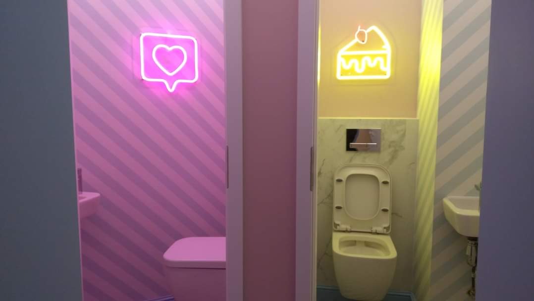 Dwa wejścia do łazienek z neonowymi dekoracjami: różowa łazienka z sercem w dymku i niebiesko-żółta z kawałkiem ciasta, każda z toaletą i umywalką w dopasowanych kolorach.