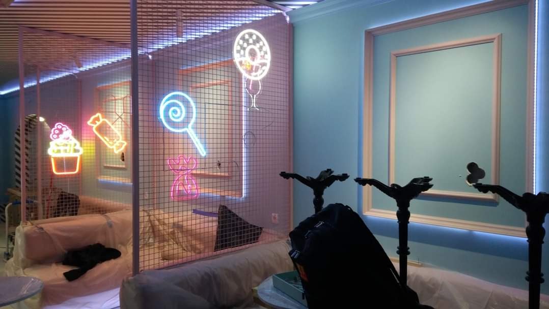 Wnętrze kawiarni lub cukierni w trakcie aranżacji, z neonowymi dekoracjami w kształcie słodyczy na tle kraty, niebieskie ściany z listwami LED, meble zabezpieczone folią ochronną, widoczne elementy...