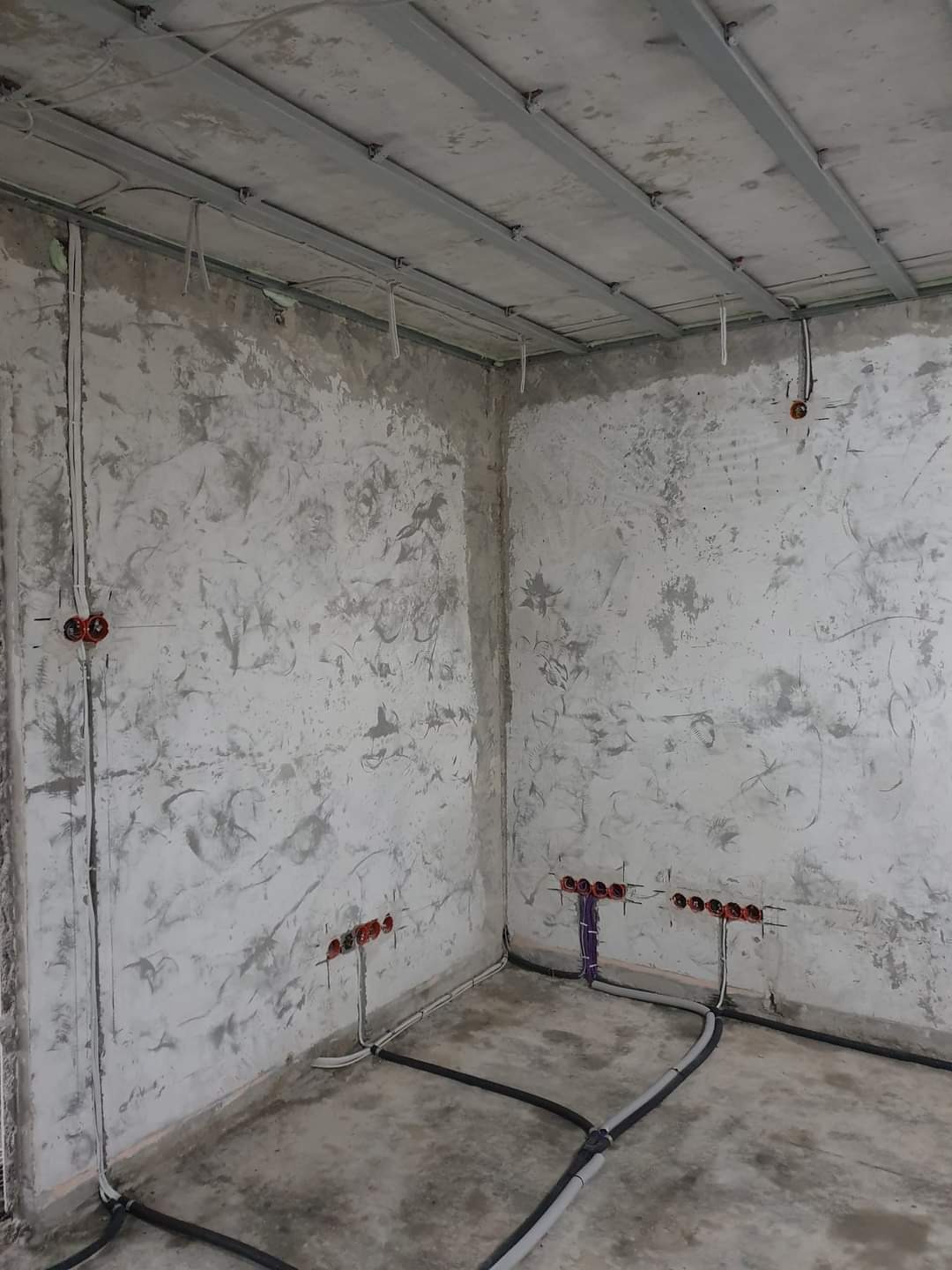 Surowy stan wnętrza w trakcie prac: widoczne przygotowane punkty elektryczne w ścianach o szorstkiej fakturze, metalowa konstrukcja sufitu podwieszanego i kable prowadzone po podłodze.