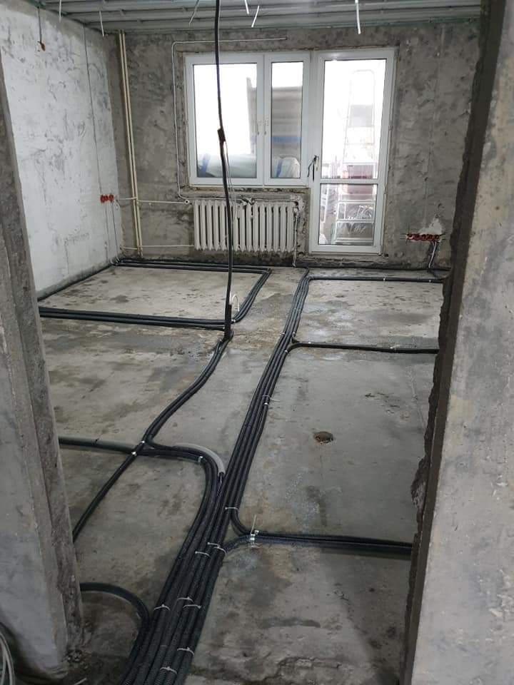 Surowy stan wnętrza w trakcie rozprowadzania instalacji elektrycznej w peszlach na podłodze, widoczne kable wychodzące ze ściany i okno balkonowe.