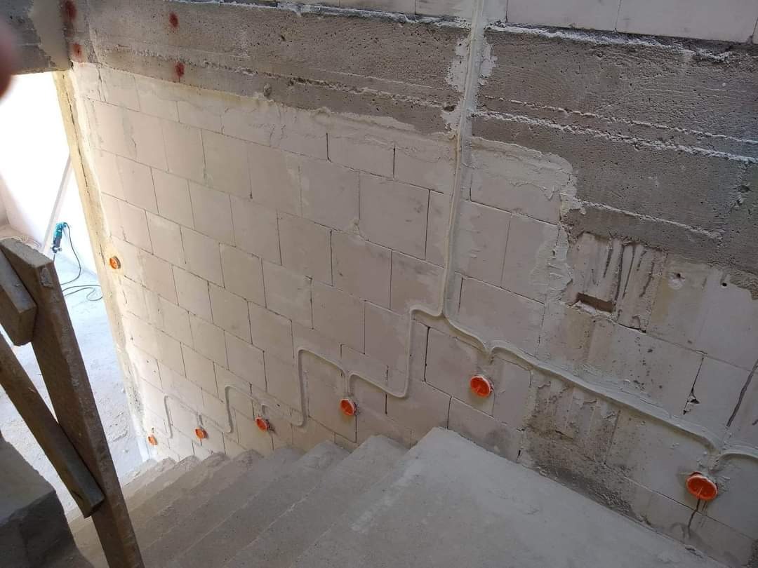 Wewnętrzna ściana schodów w trakcie instalacji elektrycznej: widoczne przewody w listwach i pomarańczowe puszki elektryczne, ściana z betonu komórkowego, częściowo otynkowana.