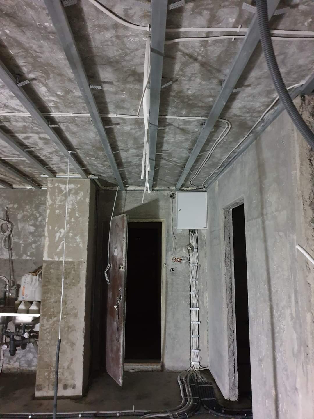 Surowy stan wnętrza w trakcie prac: widoczne metalowe profile pod sufitem, okablowanie elektryczne prowadzone po ścianie, otwarta metalowa brama, surowe betonowe ściany i podłoga.
