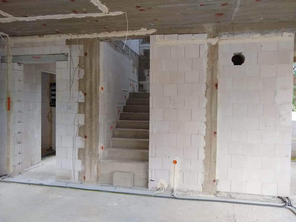 Wnętrze w stanie surowym: ściany z białych bloczków, betonowe schody, widoczne kable elektryczne i otwory instalacyjne, sufit w trakcie wykończenia.