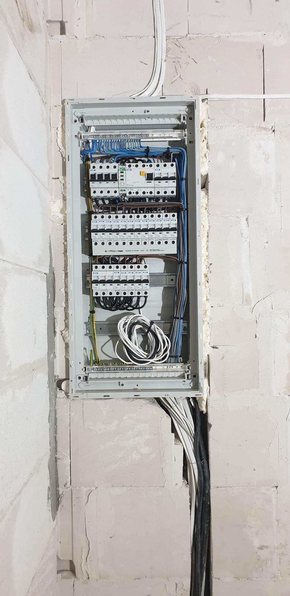 Otwarta skrzynka elektryczna z zainstalowanymi bezpiecznikami i podłączonymi przewodami w surowej ścianie z betonu komórkowego, zabezpieczona pianką montażową.